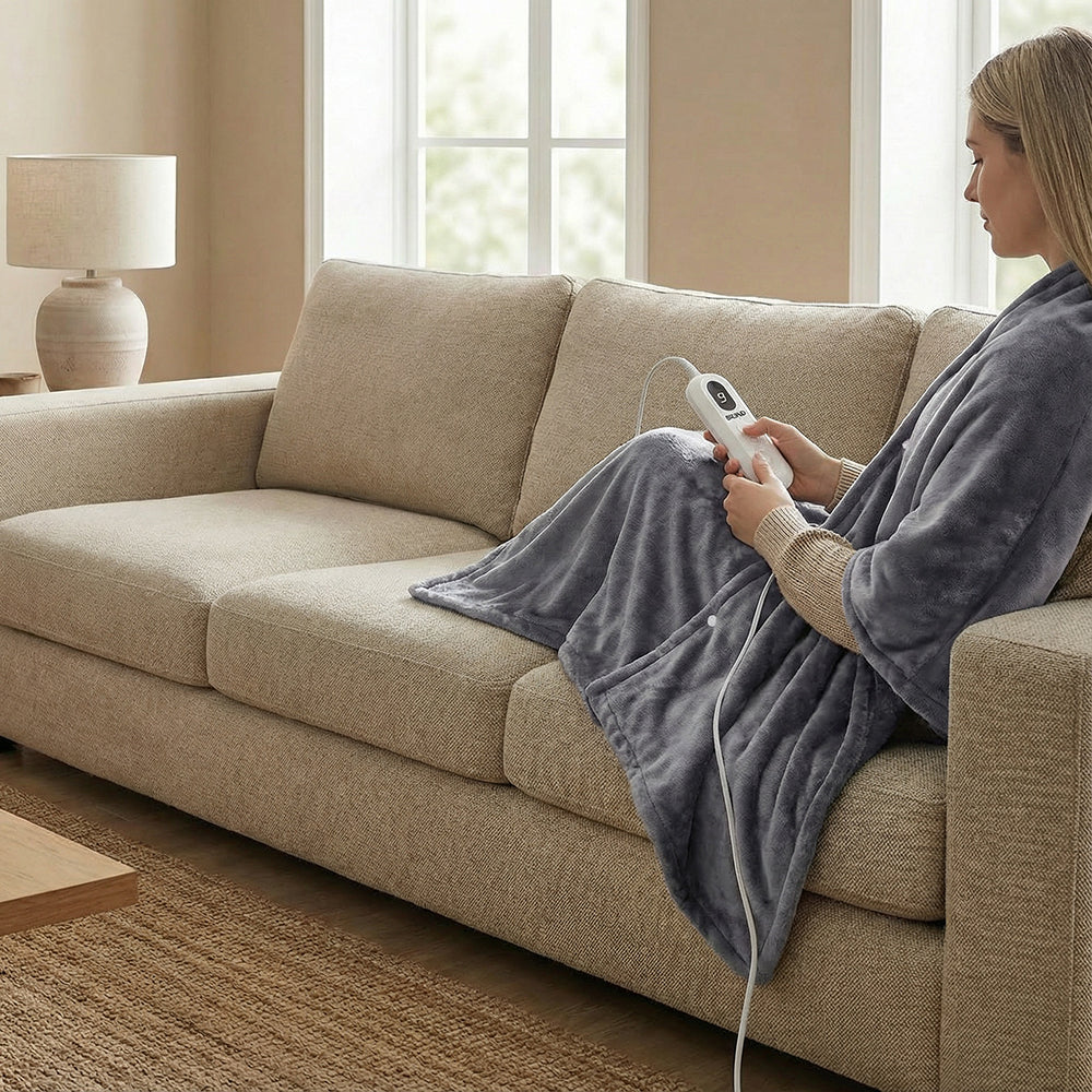 Une femme assise sur le canapé porte le poncho chauffant gris, la commande à la main
