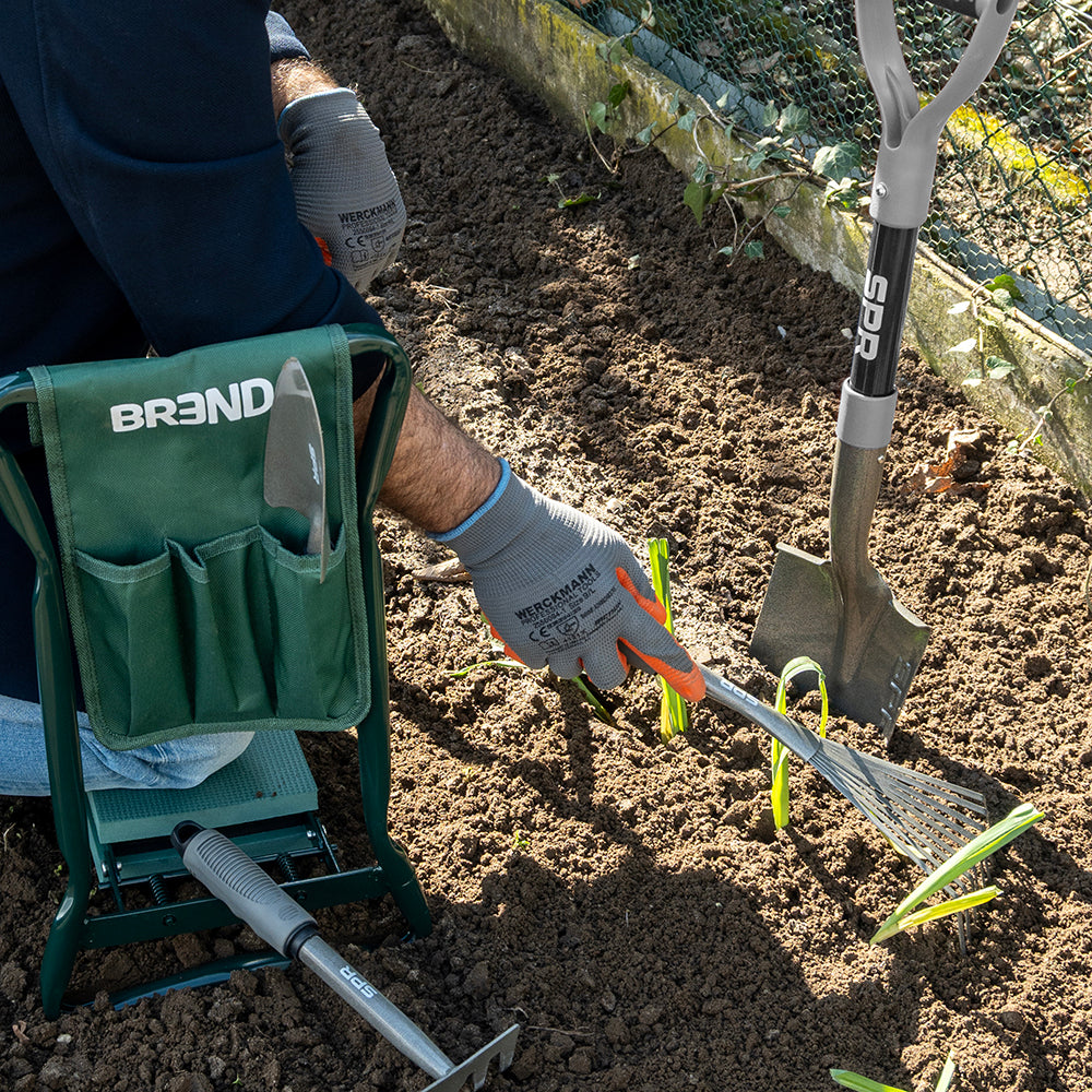 Persoon op een tuinkruk werkt met de hark tussen jonge plantjes, de spade staat in de grond