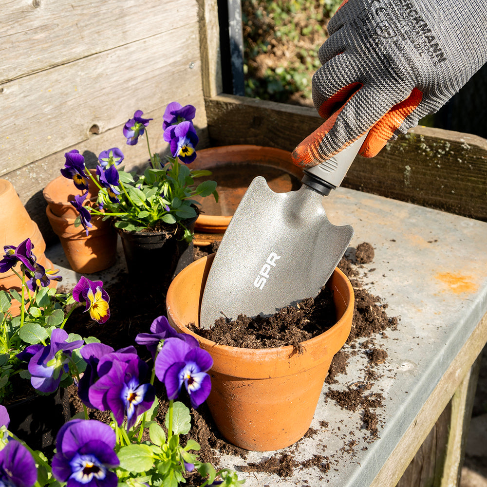 Hand met werkhandschoen schept potgrond in een bloempot met viooltjes met de brede schep